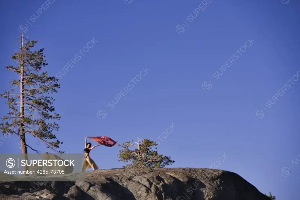 A dancer with a blowing scarf on top of a rock next to boulders and pine trees in the Sierra mountains near Truckee California