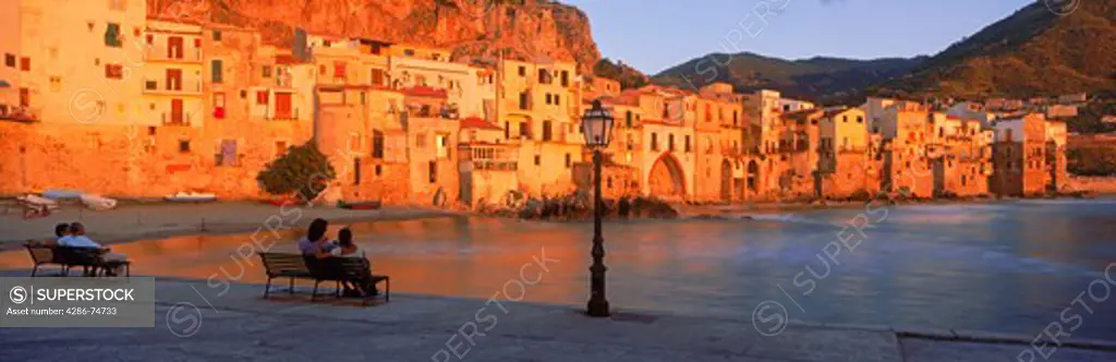 Couples on waterfront benches at sunset in village of Cefalu on Sicily