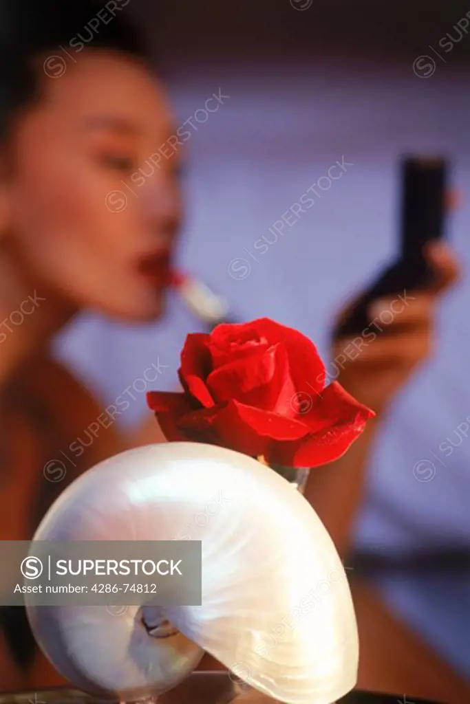 Lady applying lipstick in bedroom behind nautilus shell and red rose