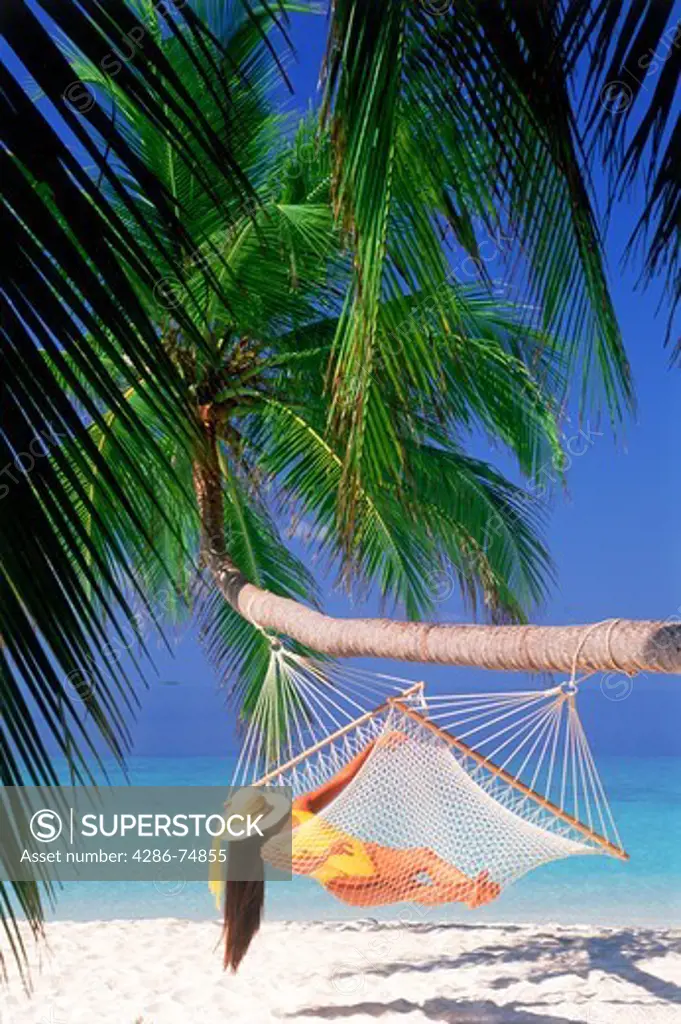 Woman with hat and yellow bathing suit relaxing in hammock under green palms over white sandy beach
