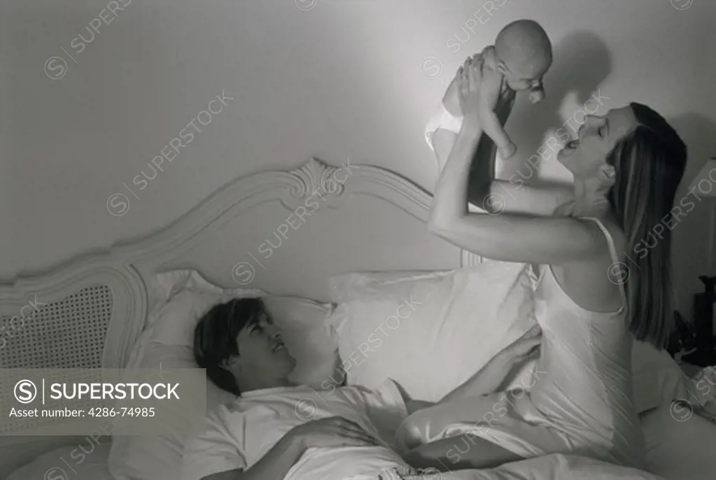 Mother holding baby overhead in bed above proud father