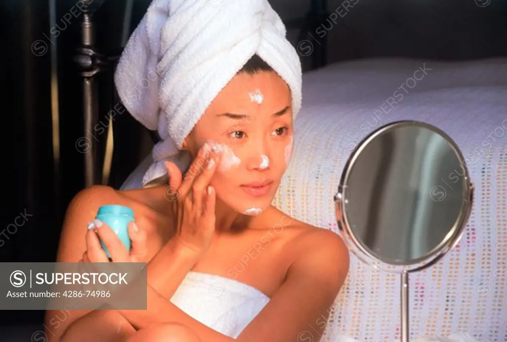 Asian woman applying moisture creme to face using small mirror in bedroom