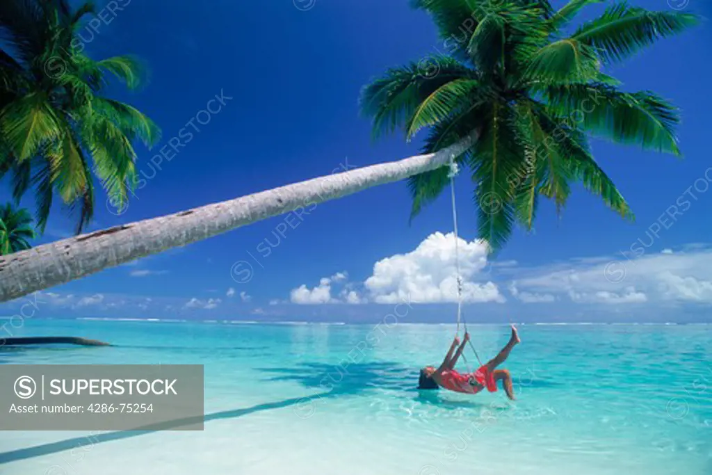 Woman laying in swing over aqua waters under palm tree and clouds