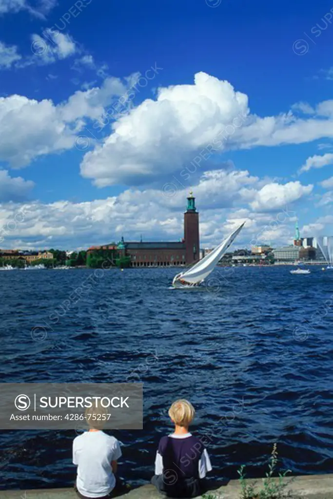 Two boys sitting on waters of Riddarfjarden with sailboat and City Hall in Stockholm