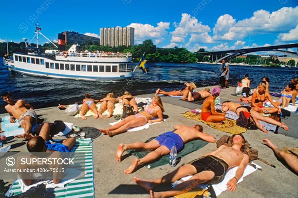 Sunbathers on cement pontoon at Langholmen Island in Stockholm with passing ferryboat on Riddarfjarden