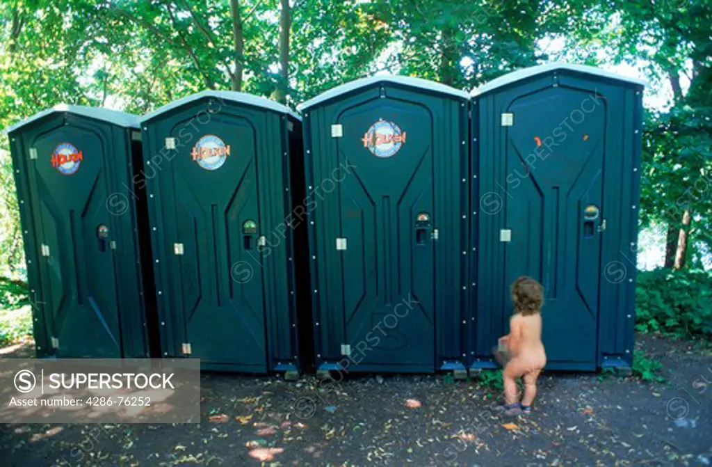 Yound child waiting naked outside line of outhouses or portable toilets at Stockholm city beach park in summer
