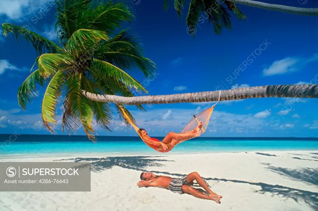 Couple chilling out with hammock under palm trees and sunny skies on white sandy beach in Maldives