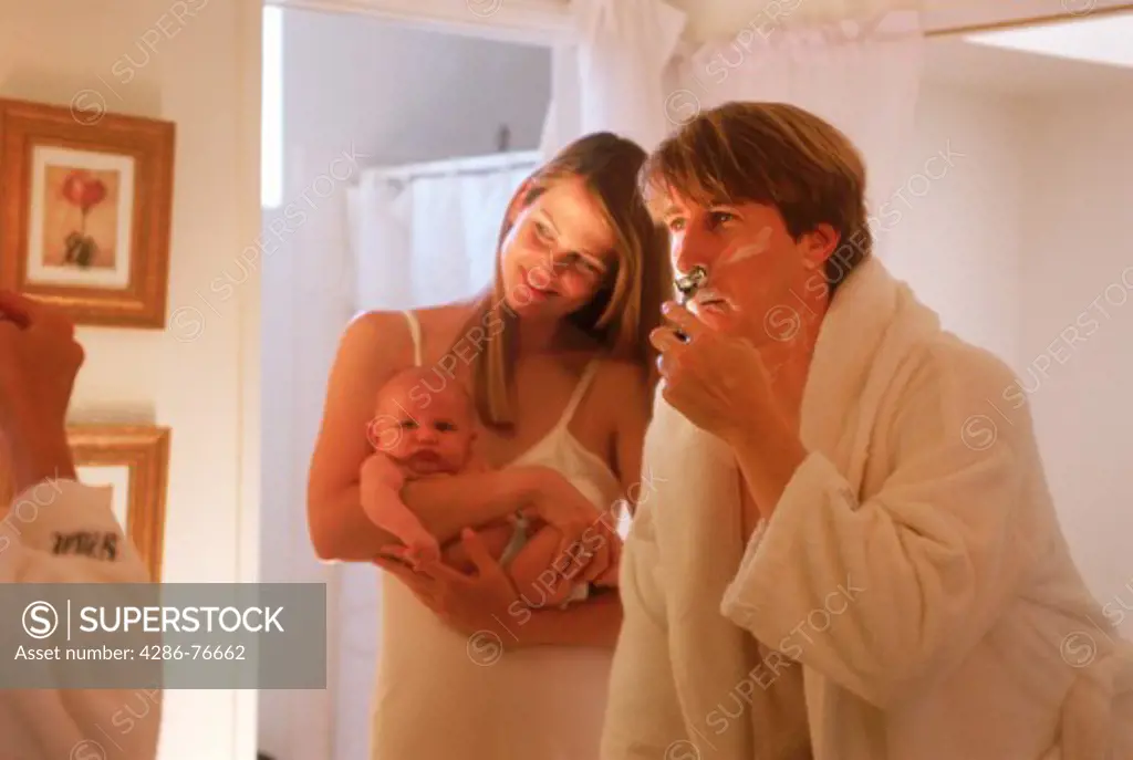 Dad shaving at home with wife holding baby in bathroom reflecting off mirror