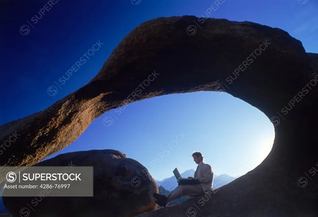 Man at outdoor office sitting under arch with computer in California Sierra Nevada Mountains within the Alabama Hills at sunrise