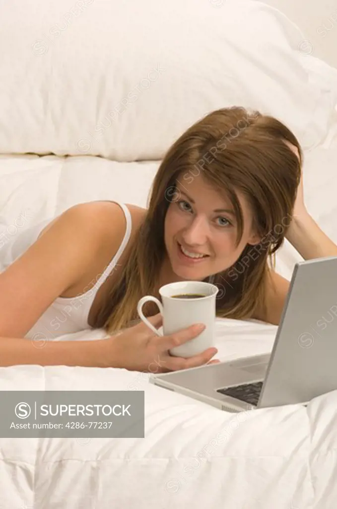 Young woman using her laptop computer while reclining in bed and drinking coffee