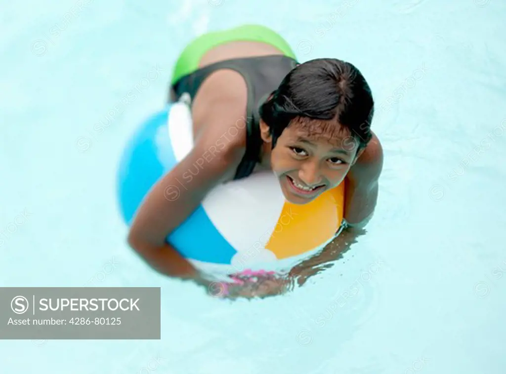 A nine year old girl plays on a beach ball in a swimming pool. 