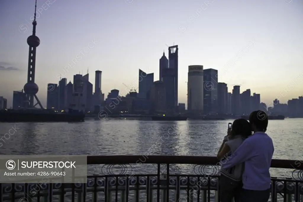 Young couple take photo with cellular phone and cuddle before dawn along skyline, The Bund, Shanghai, China