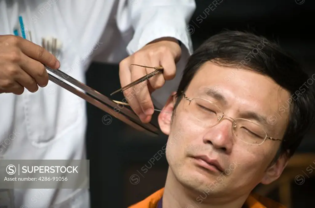 Outdoor ear-cleaner stimulates man's acupressure point with feather brush and tuning fork, Jin Li Street, Chengdu, Sichuan Province, China