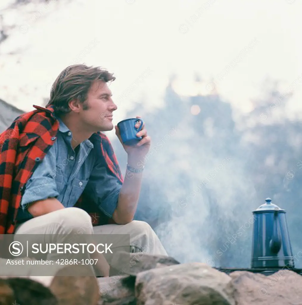 Rugged outdoors man drinking a cup of coffee by his campfire with his flannel shirt draped over his shoulders.