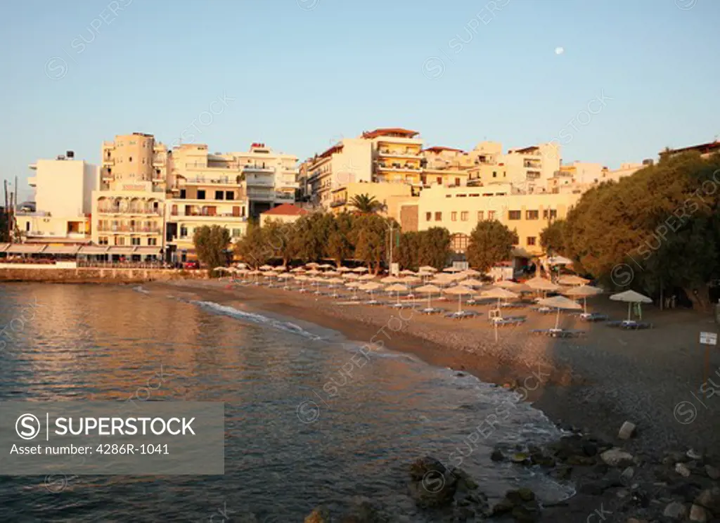 An inviting beach at dawn in Aghios Nikolaos, Crete, with its sunbeds ready to receive the day's holidaymakers. The moon is visible in the sky.