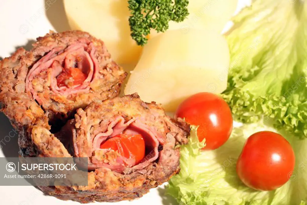 Slices of beef roulade (rolled beef meatloaf) served with potato, lettuce and cherry tomato. The roulade is wrapped around ham and tomatoes.