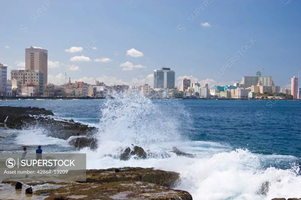 Boys play on the rocks as waves crash along the Malecon in Old Havana, hotels of Vedado are in the distance