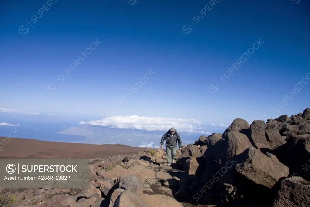 Man hiking through lava boulders on the 10,000 foot Haleakala volcano on Maui. West Maui can be seen off in the distance.