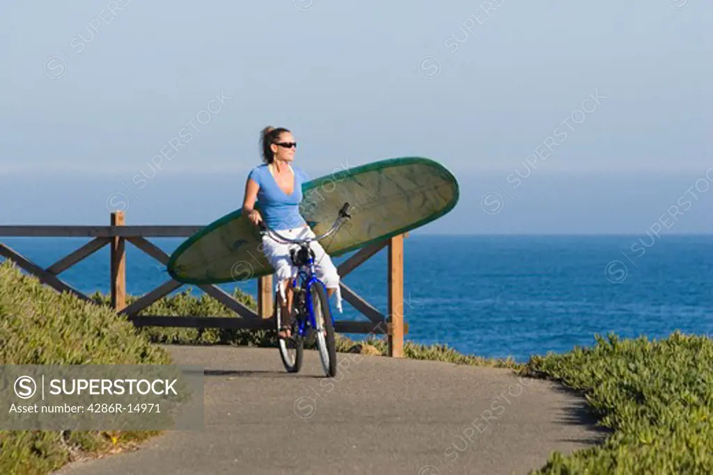A woman riding a bike with a surfboard above the ocean in Santa Cruz California