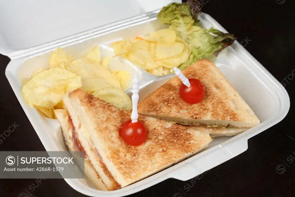 A club sandwich with lettuce and potato chips in a polystyrene box on a dark wooden table.