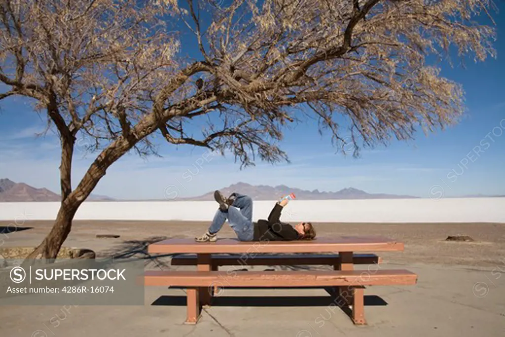 A woman lying on a picnic table reading a book under a Mesquite tree at a rest stop on highway I-80 on the Bonneville Salt Flats in Utah
