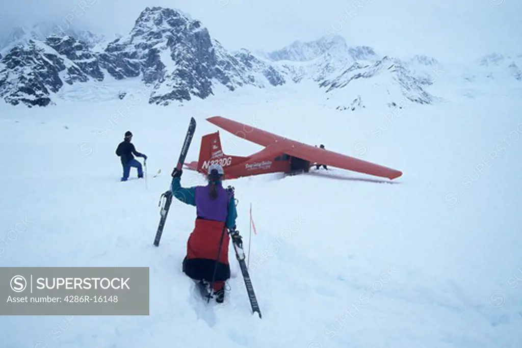 Three people and a red bush plane with skis stuck in deep snow in the Ruth Gorge in Denali National park in Alaska