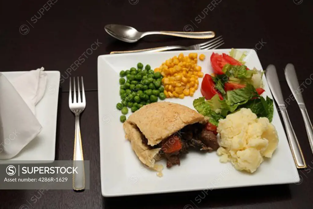 A steak pudding served with vegetables and salad on a dark wooden table.