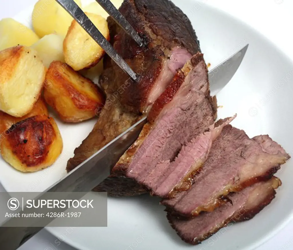 Carving a joint of roast beef on a plate with roasted and boiled potatoes.