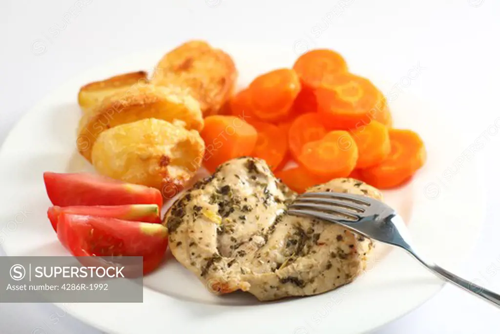 A chicken breast, marinated with oregano and then baked, served with roasted potatoes, boiled carrots and sliced tomato