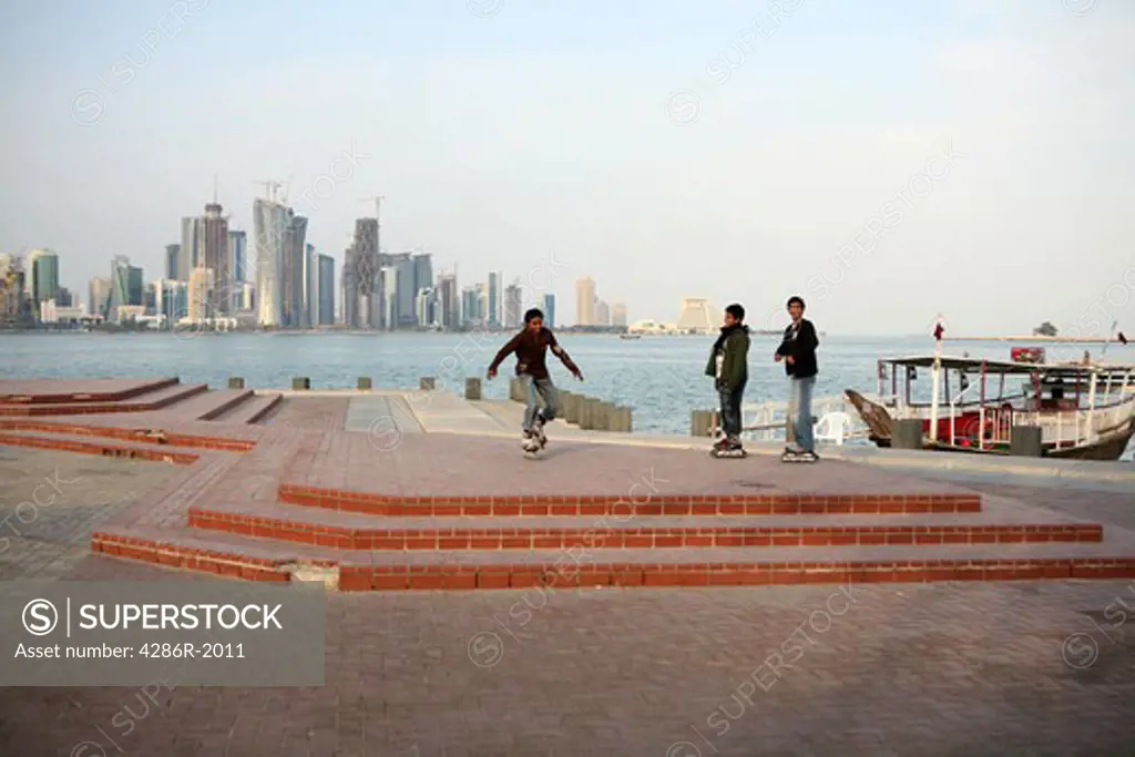 A group of boys amuse themselves rollerblading on the Corniche in Doha, Qatar, with the new high-rise West Bay district skyline visible behind them