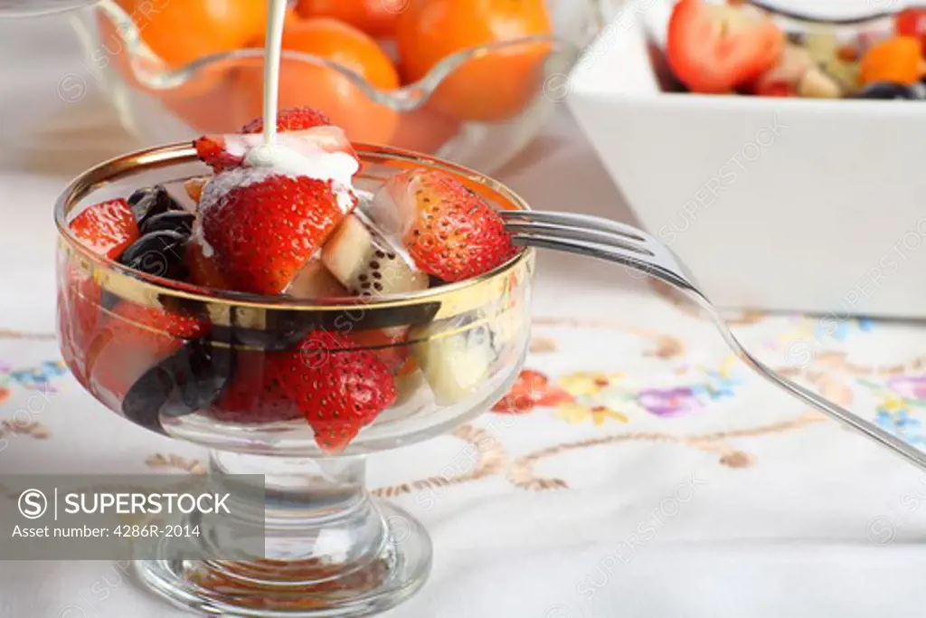 A glass dessert bowl with fresh fruit salad in it and cream being poured over