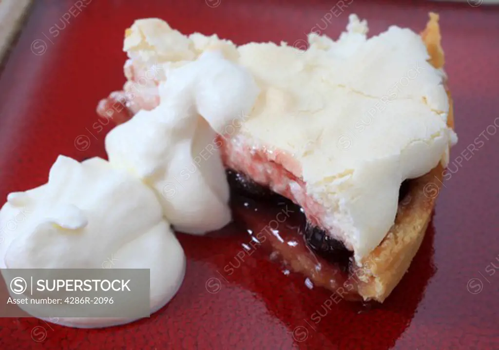 A slice of meringue pie with a cherry filling on a red plate with dollops of whipped cream