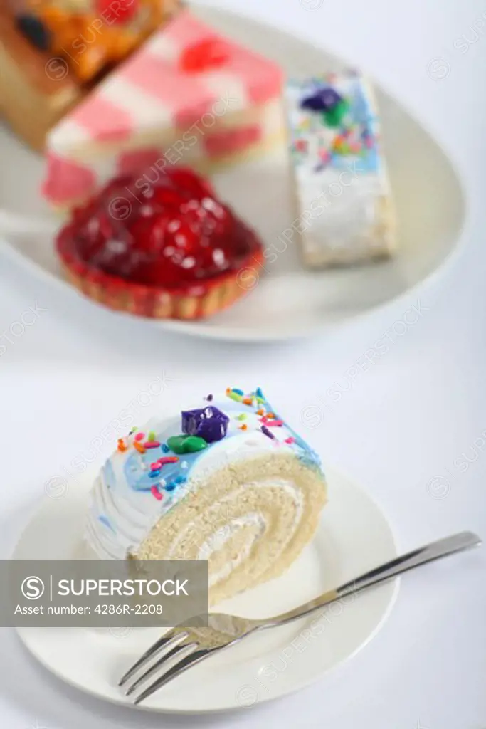 A colourful sponge roll with a fork and a plate of other cakes in the background