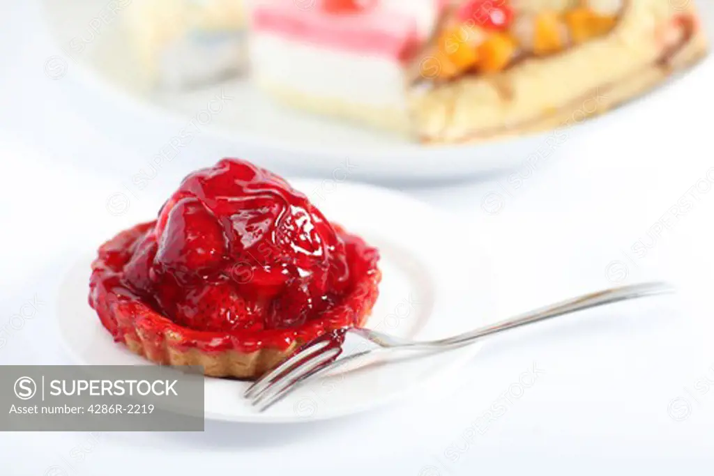 A strawberry tart and fork with a plate of other cakes in the background