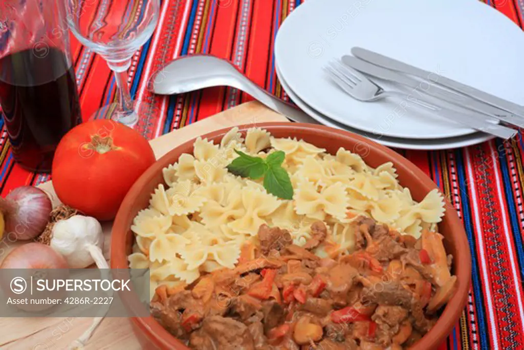 A terracotta serving bowl with beef stroganoff and pasta bows, with plates, cutlery, and ingredients on a bright tablecloth