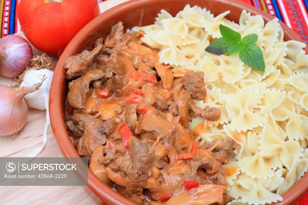 Close-up view of a terracotta bowl of beef stroganoff and pasta bows, with a chopping board and ingredients