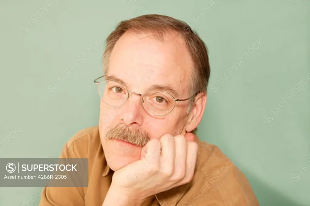 Studio shot of a middle aged man with a mustache and his chin in his hand wearing glasses.