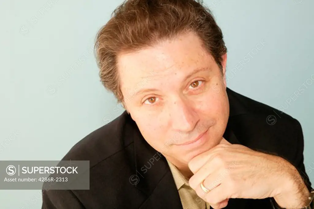 Studio shot of a brown-haired man with his chin on his hand and he has a gloomy look on his face.