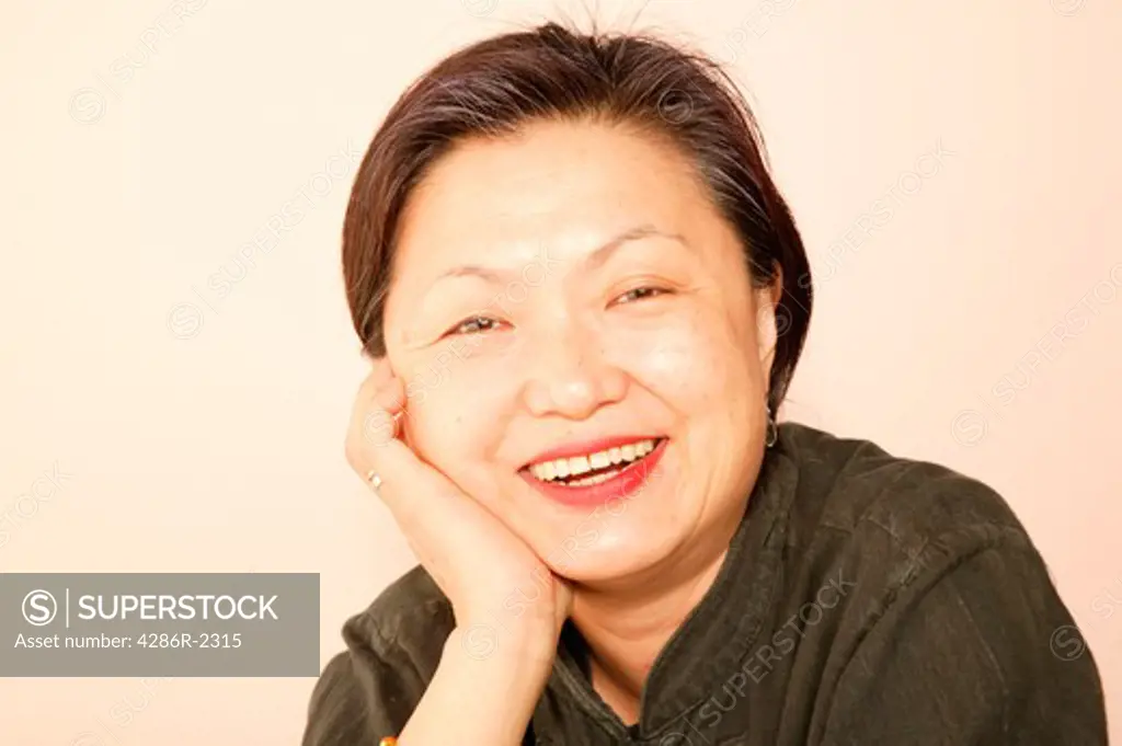 Studio shot of an Asian woman with her cheek in her hand smiling at the camera.