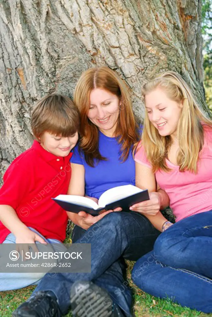 Family of mother and children reading a book under a tree in summer park