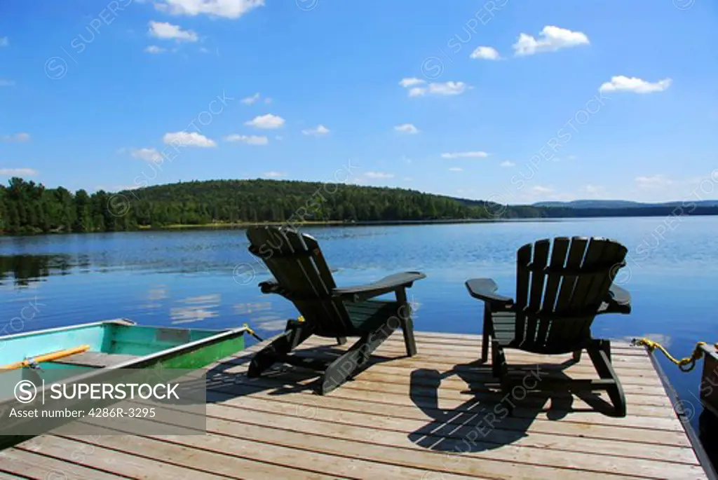 Two adirondack wooden chairs on dock facing a blue lake with clouds reflections