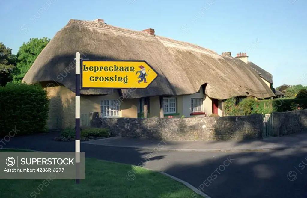 A thatched cottage along a country road in Ireland with a road sign for  Leprechaun crossing posted in foreground.