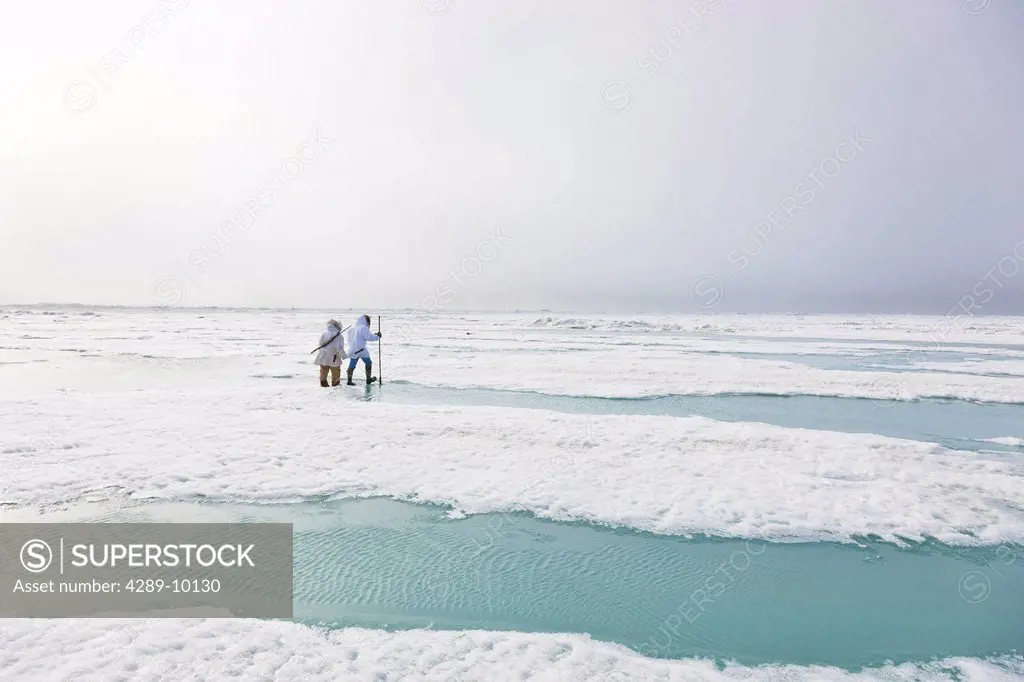 Two Inupiaq Eskimo hunters wearing their Eskimo parka´s Atigi carry a rifle and walking stick while walking over the shore ice along the Chukchi Sea, ...