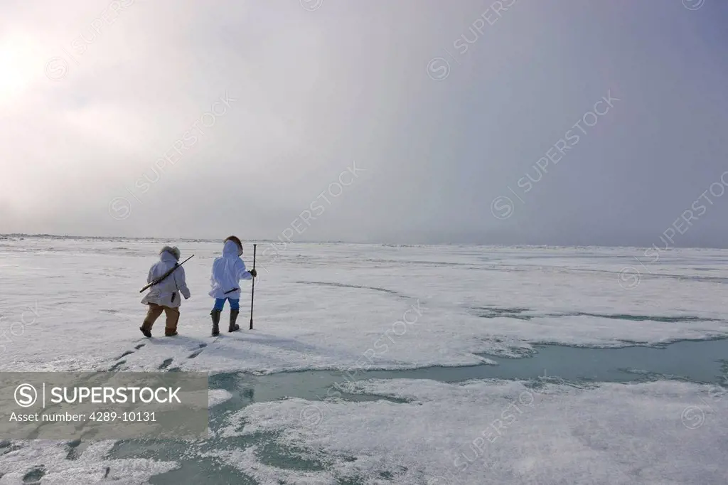 Two Inupiaq Eskimo hunters wearing their Eskimo parka´s Atigi carry a rifle and walking stick while walking over the shore ice along the Chukchi Sea, ...