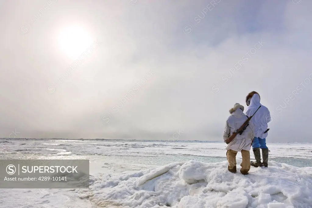 Male and female Inupiaq Eskimo hunters wearing their Eskimo parka´s Atigi carry a rifle and walking stick while looking out over the Chukchi Sea, Barr...