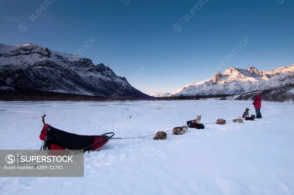 National Park Ranger rests with his dog team on the North Fork of the Koyukuk River at sunset with the Gates of the Arctic in the background, Brooks R...