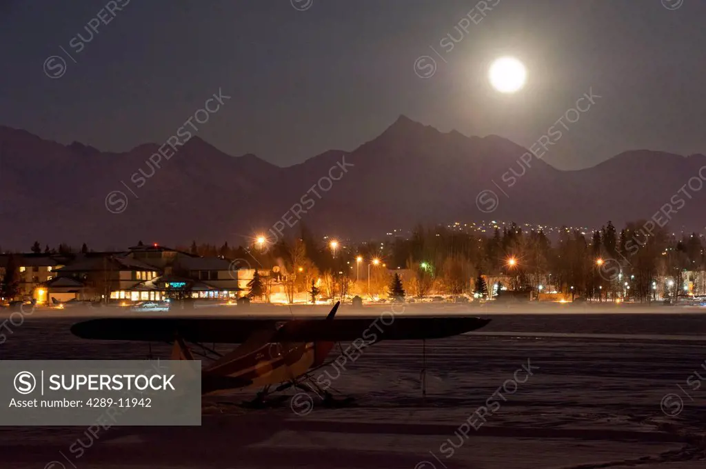 A full moon rises over the Chugach Mountains with a ski plane parked on the frozen Lake Hood in the foreground, Southcentral Alaska, Winter