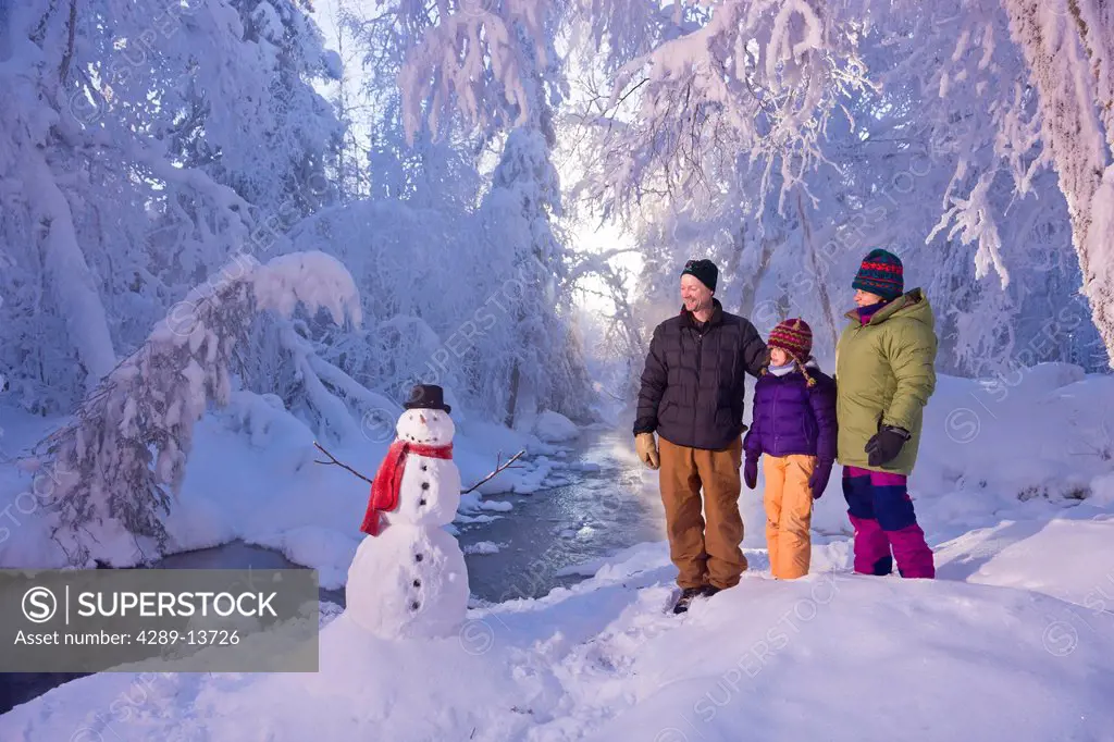 Family stands next to a snowman and small stream in a hoarfrost covered forest, Russian Jack Springs Park, Anchorage, Southcentral Alaska, Winter