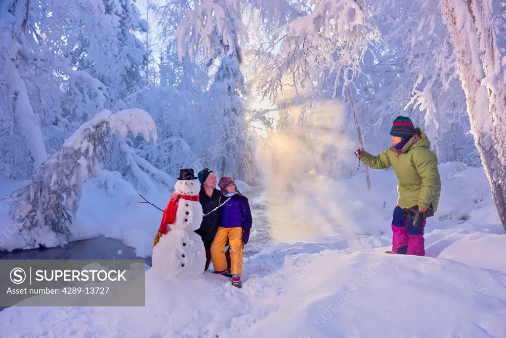 Mother photographs husband and daughter posing next to a snowman in a frost covered forest, Russian Jack Springs Park, Anchorage, Southcentral Alaska,...