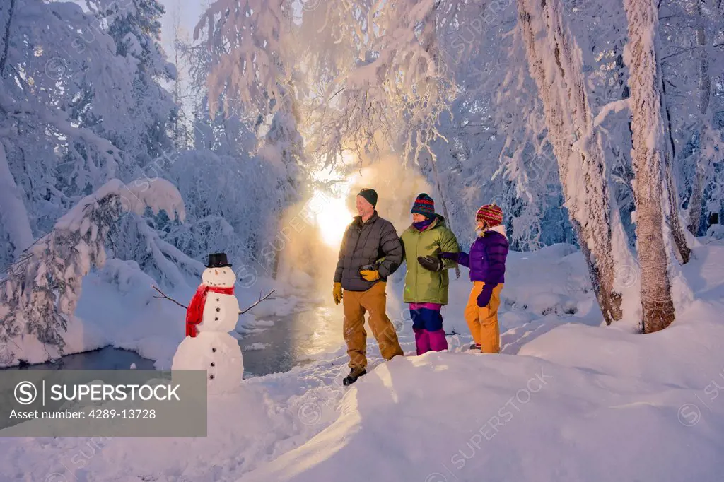 Family stands next to a snowman and small stream in a hoarfrost covered forest, Russian Jack Springs Park, Anchorage, Southcentral Alaska, Winter